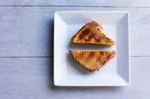 Top-down view of a toasted ham and cheese sandwich cut in half on a white square plate.