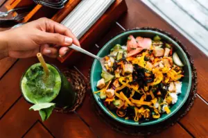A person holding a spoon over a colorful vegetarian poke bowl with tofu and vegetables, served alongside a refreshing green juice.