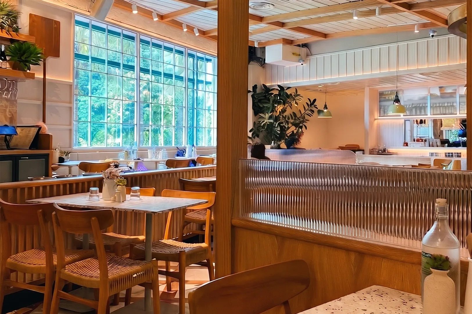 Bright modern cafe interior with stylish wooden dining tables, woven chairs, and a large grid window overlooking green trees.