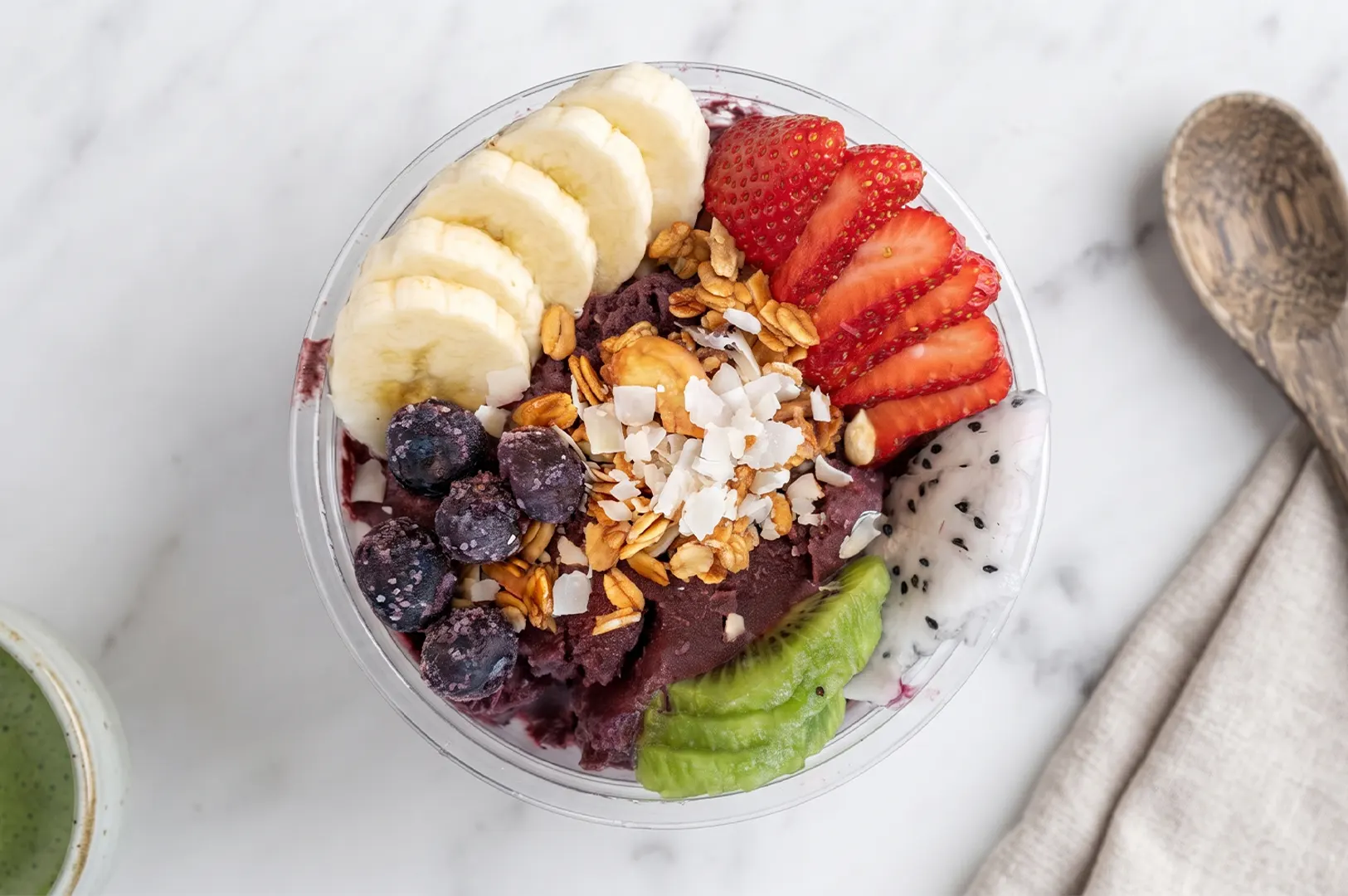 A colorful acai bowl topped with sliced bananas, strawberries, blueberries, kiwi, dragon fruit, granola, and coconut flakes.