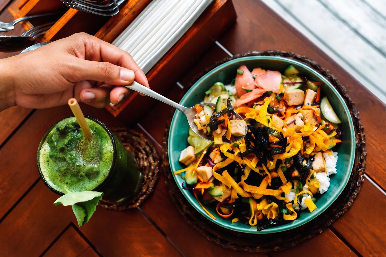 A person holding a spoon over a colorful vegetarian poke bowl with tofu and vegetables, served alongside a refreshing green juice.