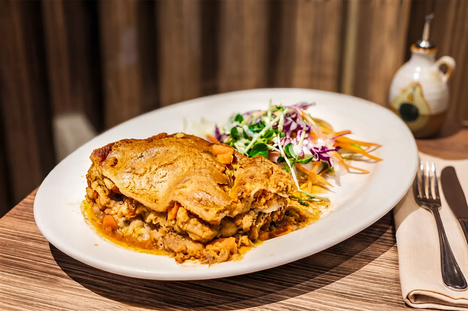 Freshly baked vegan lasagna with a crispy golden crust served with a side salad on a white oval plate, resting on a wooden table next to a cruet and cutlery.