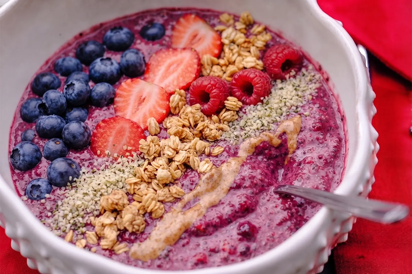 A healthy acai smoothie bowl topped with fresh blueberries, sliced strawberries, raspberries, granola, hemp seeds, and a drizzle of almond butter in a white bowl.