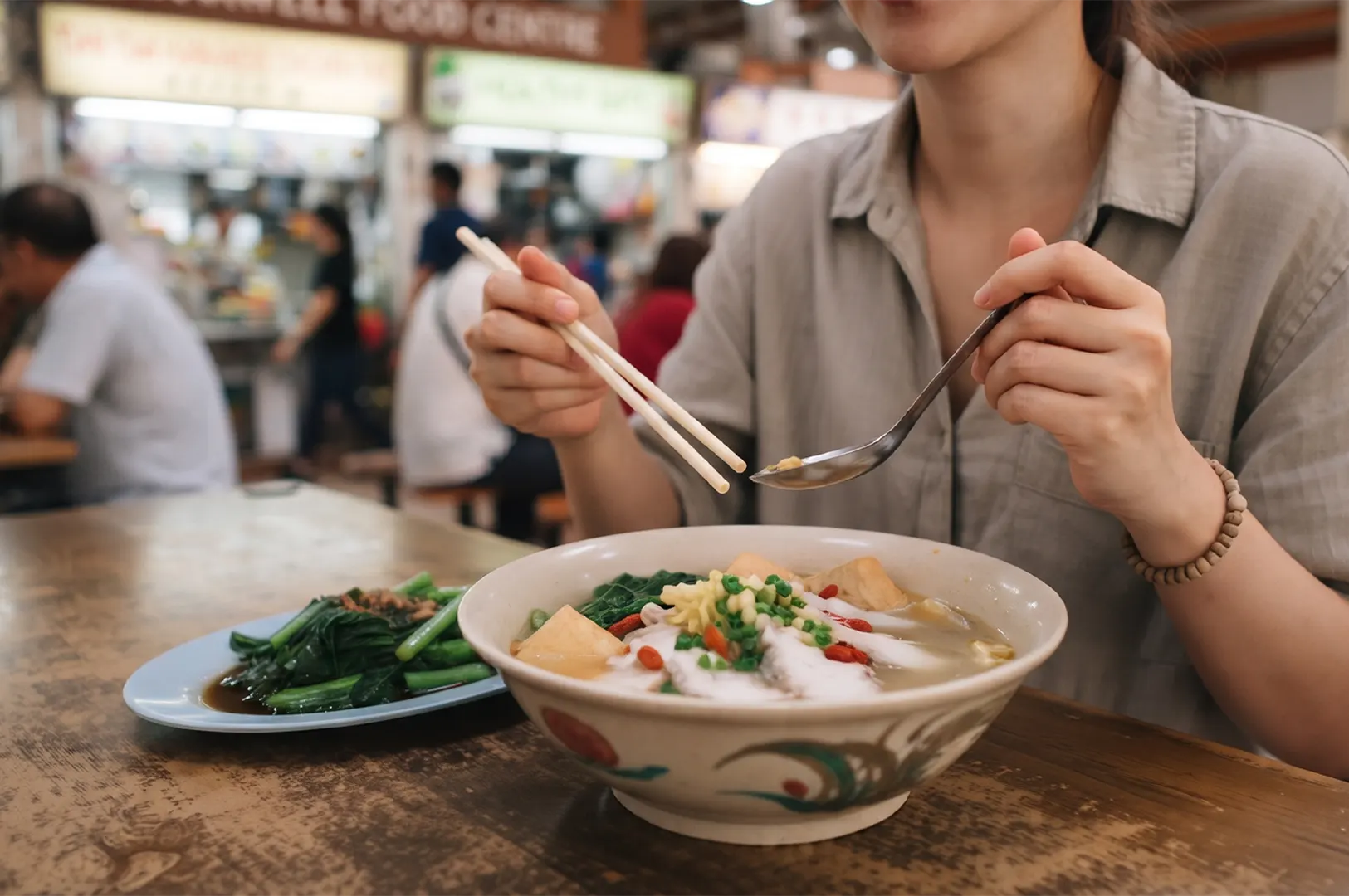 A woman eating high-protein fish soup with tofu and vegetables using chopsticks and a spoon at a food center.