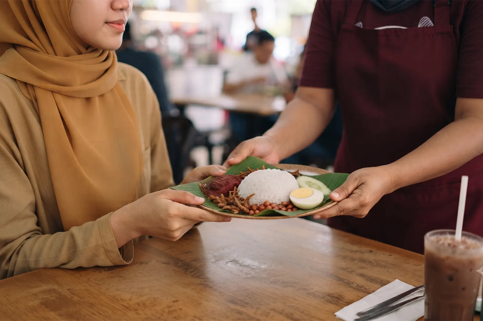 A server handing a plate of Nasi Lemak with rice, anchovies, peanuts, and egg to a customer in a restaurant.