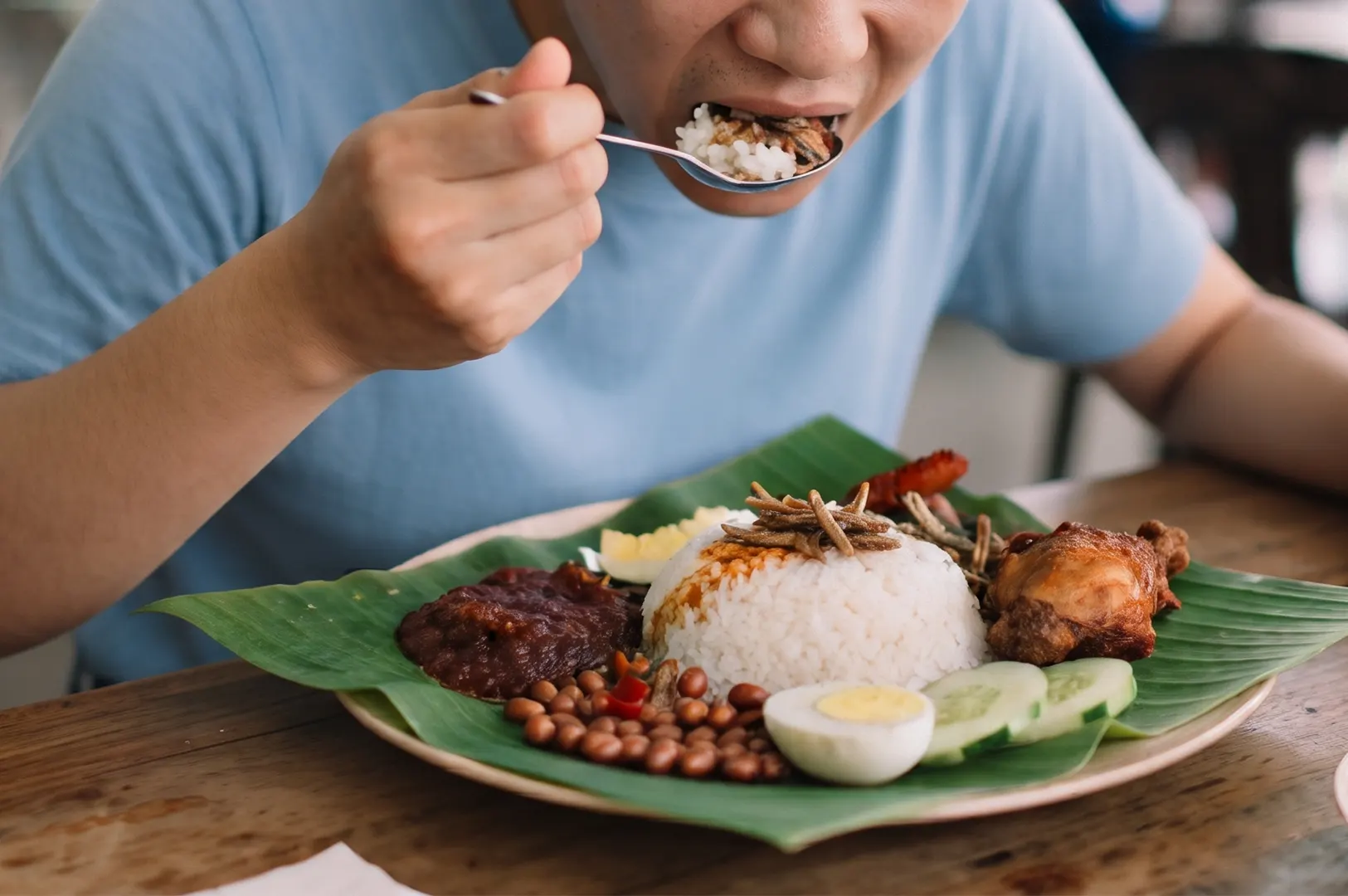 A person eating a plate of traditional Nasi Lemak with rice, fried chicken, anchovies, peanuts, cucumber, and hard-boiled egg.