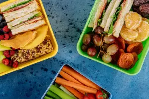 Three colorful lunch containers filled with healthy sandwiches, fresh fruits, vegetables, and snacks on a blue textured background.