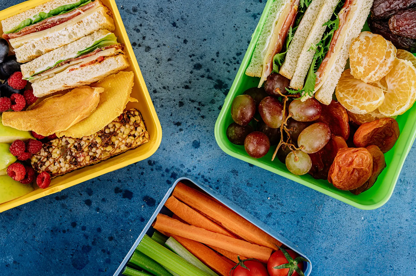 Three colorful lunch containers filled with healthy sandwiches, fresh fruits, vegetables, and snacks on a blue textured background.