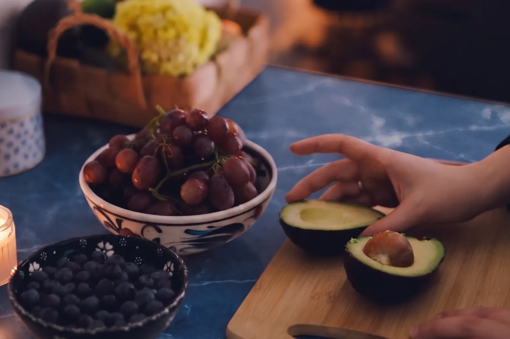 Person preparing a fresh avocado half on a wooden cutting board next to bowls of grapes and blueberries.