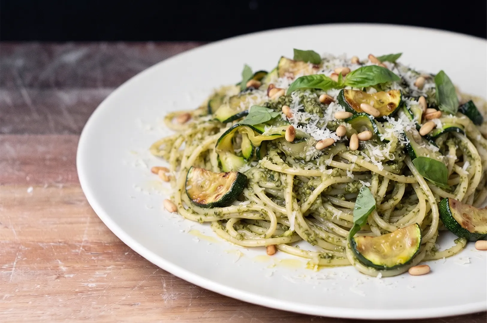 Zucchini pesto pasta with sauteed zucchini slices, toasted pine nuts, fresh basil, and grated parmesan on a white plate.