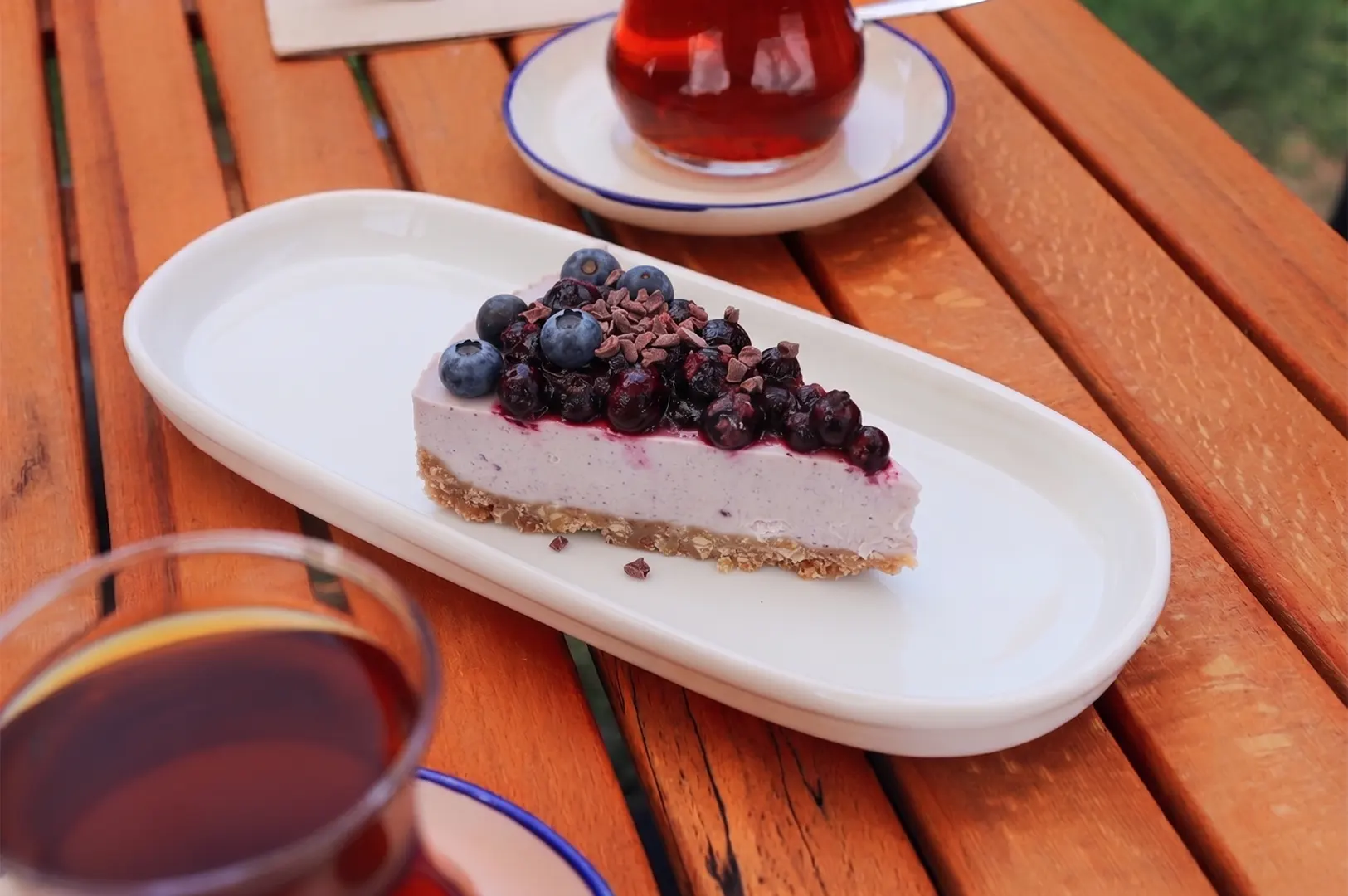 A slice of blueberry cheesecake topped with fresh blueberries and chocolate shavings, served on a white plate with tea on a wooden table.