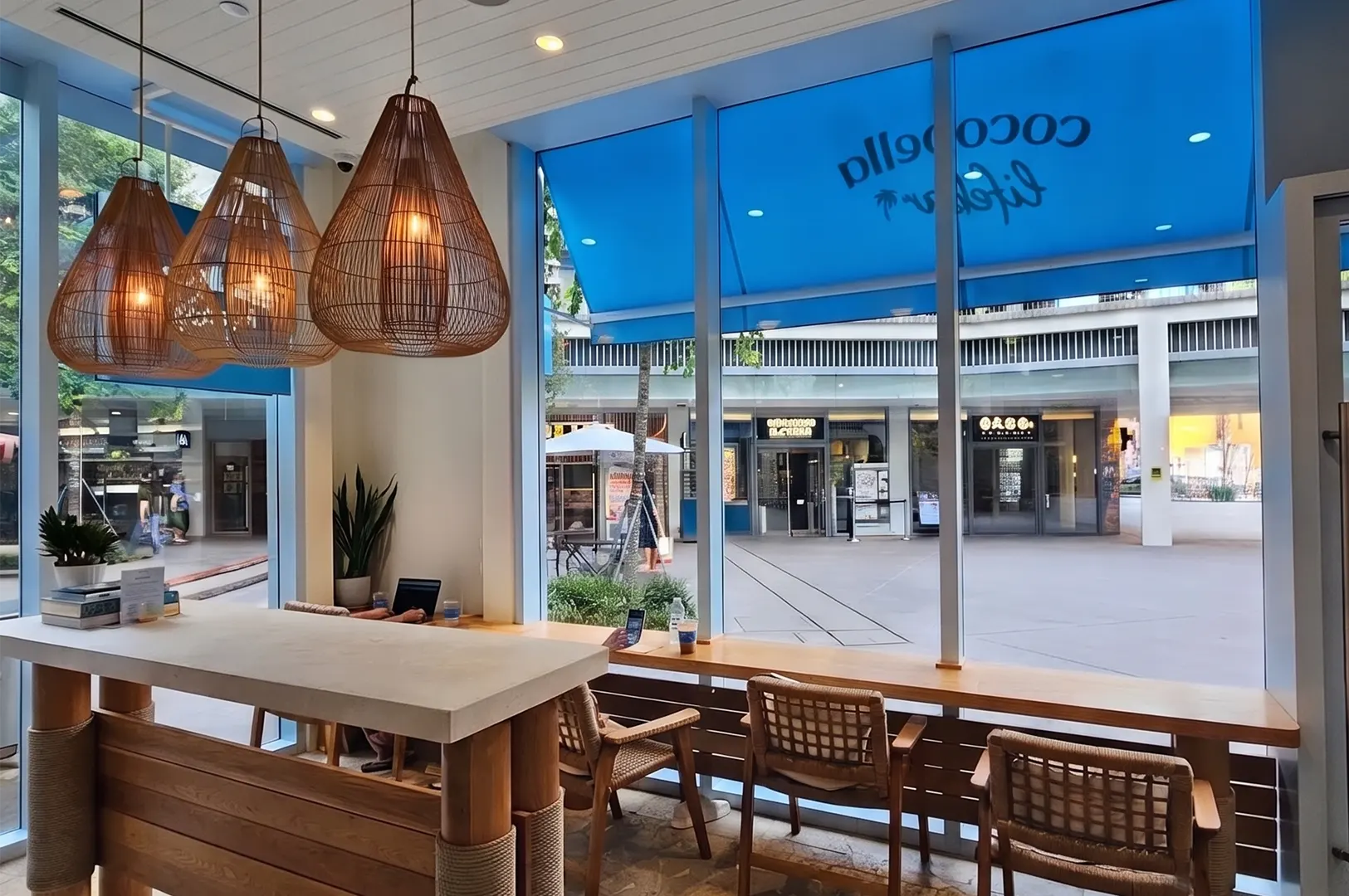 Cocobella Lifebar interior featuring modern seating, large glass windows, and woven pendant lights overlooking an outdoor plaza.