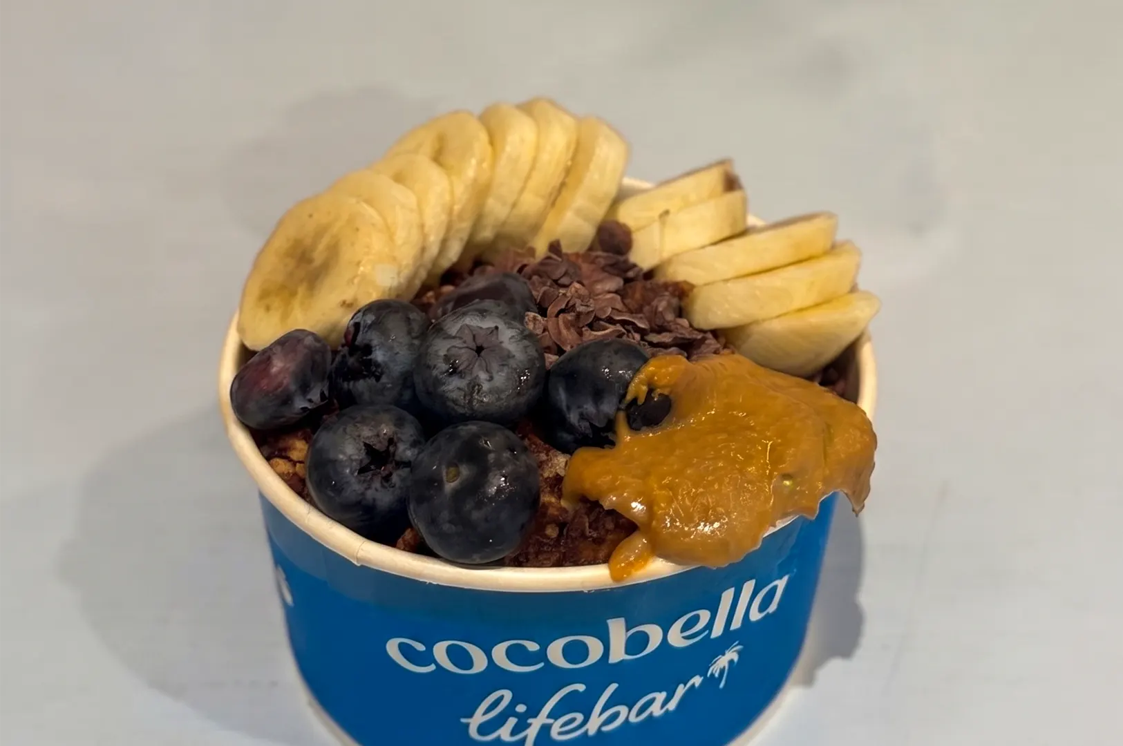 Cocobella Lifebar Choc Nut Nana yogurt bowl topped with sliced bananas, fresh blueberries, cacao nibs, and peanut butter.
