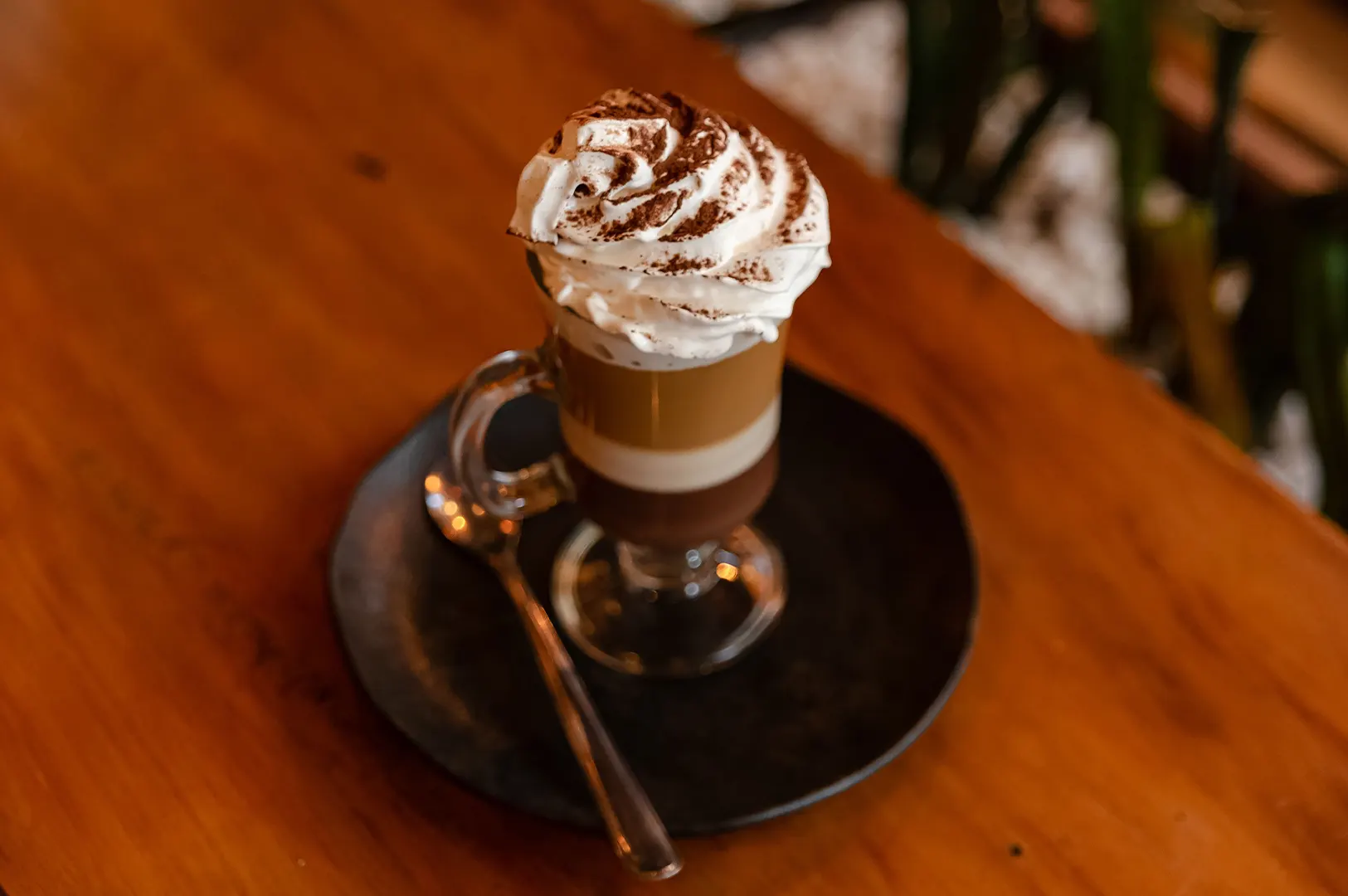 A glass of layered coffee with whipped cream and chocolate powder on a saucer.