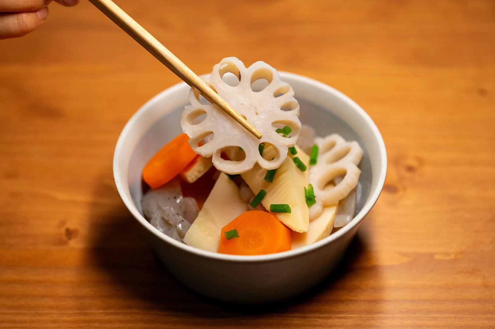 Chopsticks holding a slice of lotus root over a bowl of mixed vegetables.
