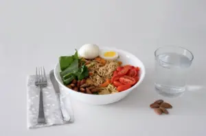 A healthy balanced meal bowl with noodles, vegetables, eggs, almonds, and a glass of water.