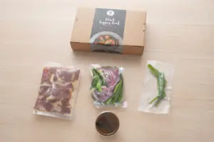 Prepped Singapore Black Pepper Beef meal kit box with fresh vacuum-packed ingredients and sauce on a wooden table.