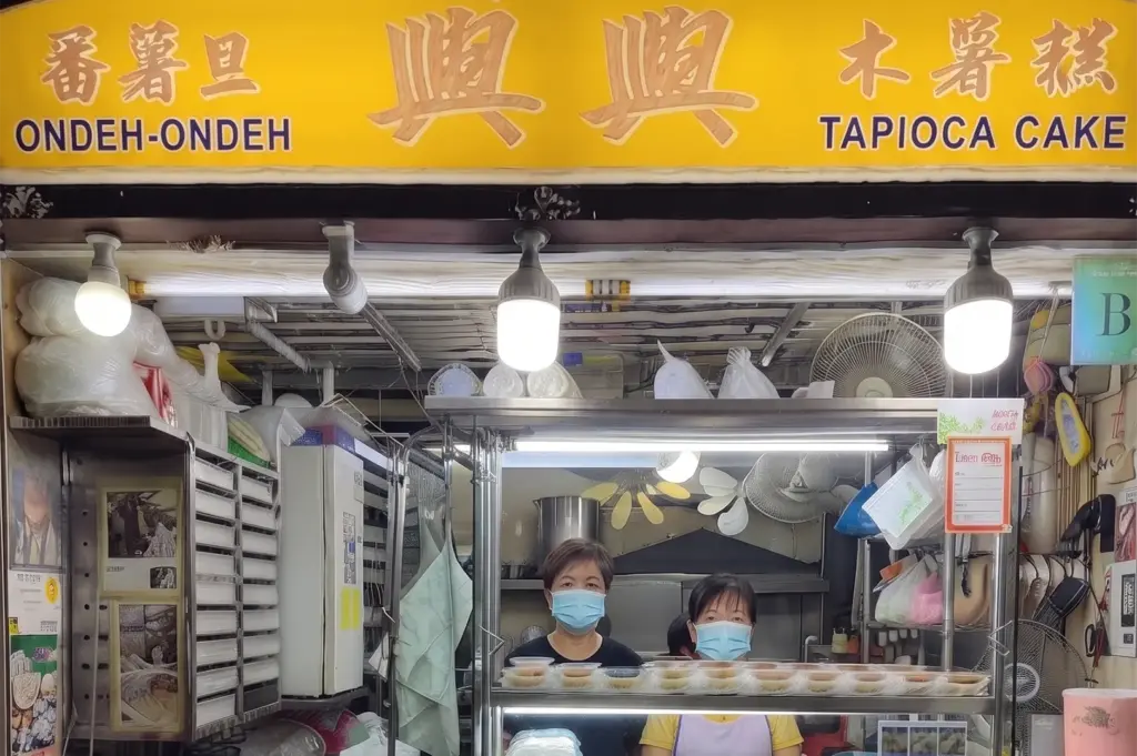 Two vendors wearing face masks working inside a traditional hawker stall selling Ondeh-Ondeh and Tapioca Cake in Singapore.