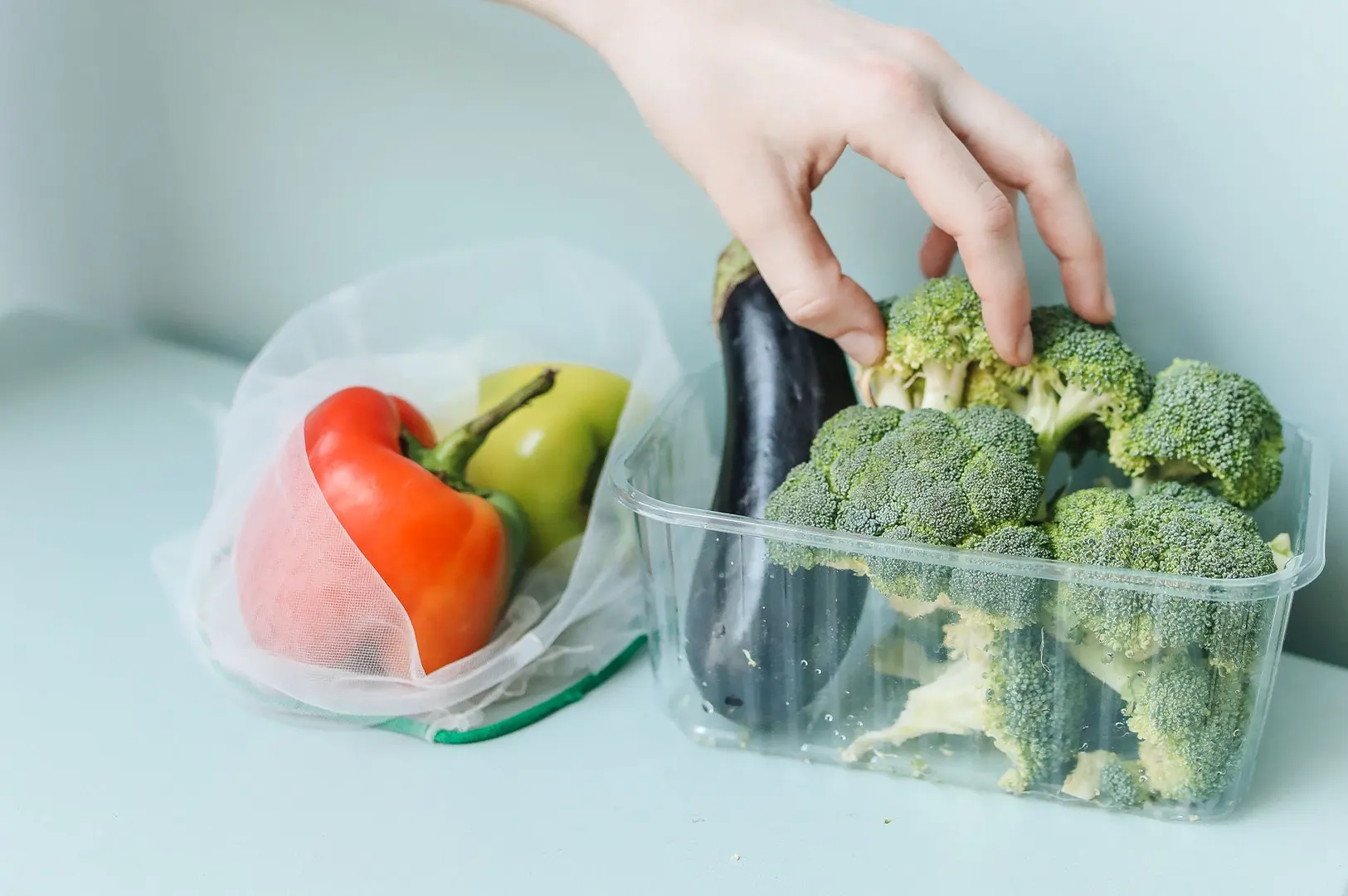 Hand picking fresh broccoli from a plastic container next to a mesh produce bag filled with bell peppers and an eggplant.
