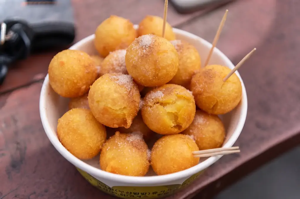 Freshly made golden sweet potato balls dusted with sugar in a paper bowl with toothpicks.