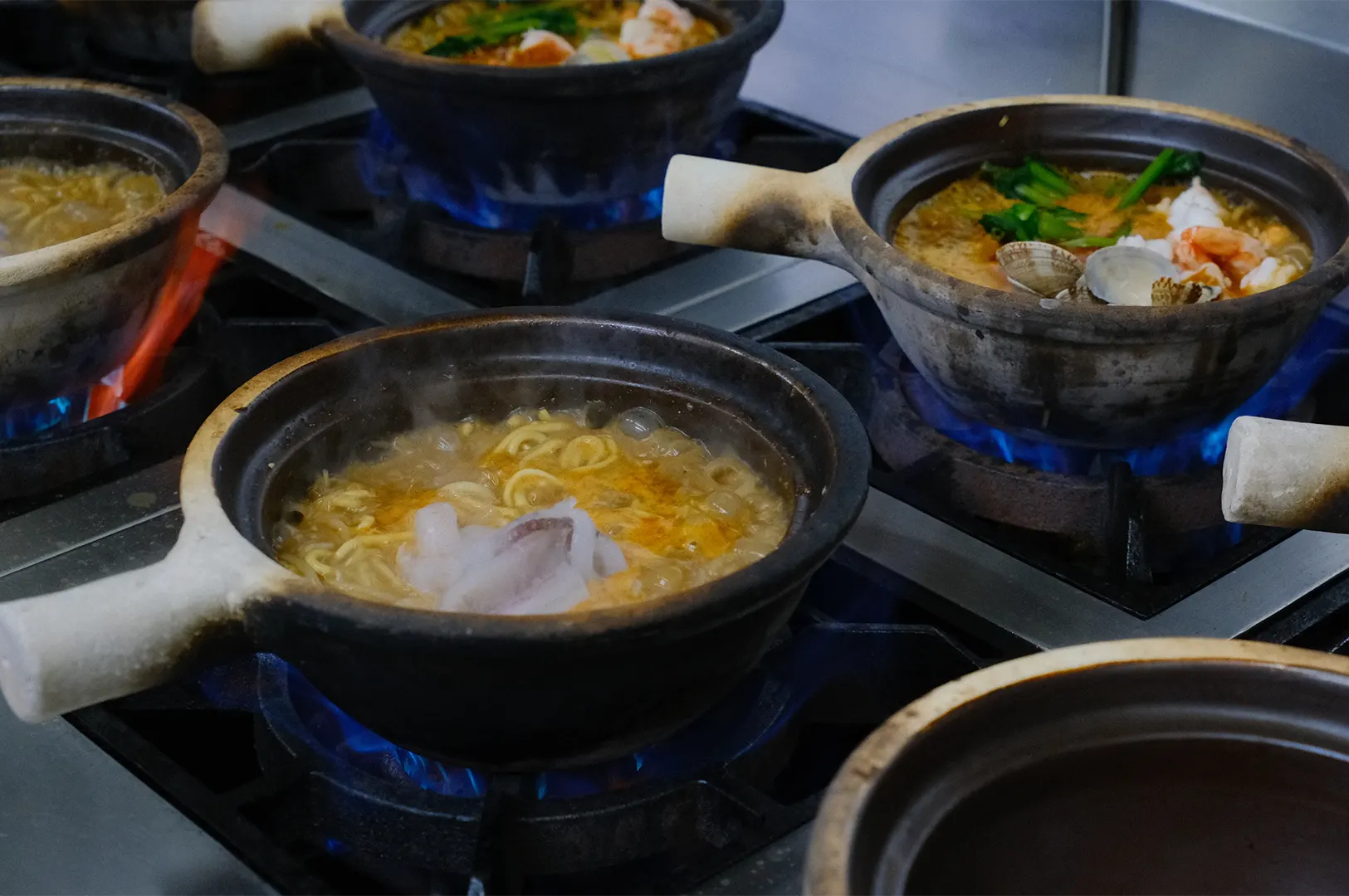 Multiple claypots cooking seafood noodles and soup on stoves.