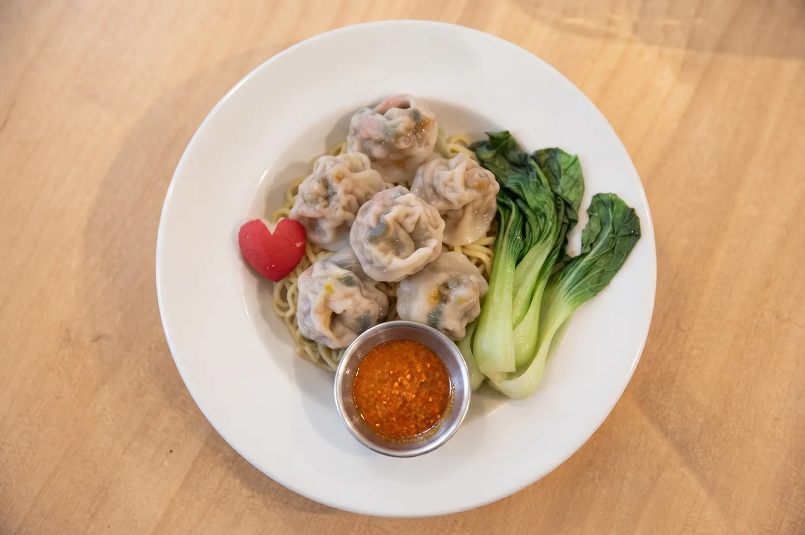 Bowl of dumpling soup with spinach noodles, tender dumplings, bok choy, and chili sauce on a wooden table.